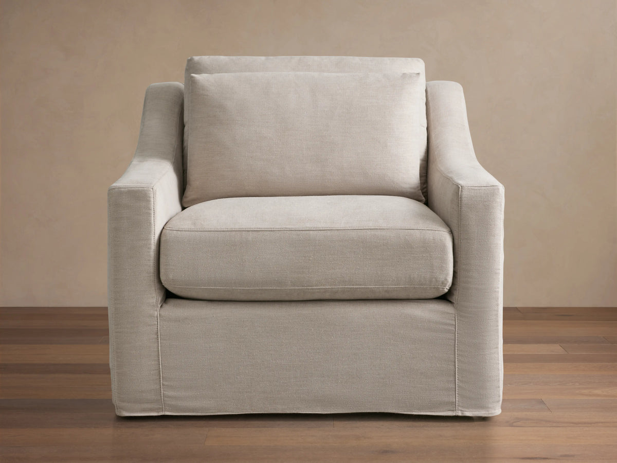 Side profile of the Ashby Slipcovered Swivel Chair in light beige fabric, showing curved backrest and plush cushions against a neutral background.