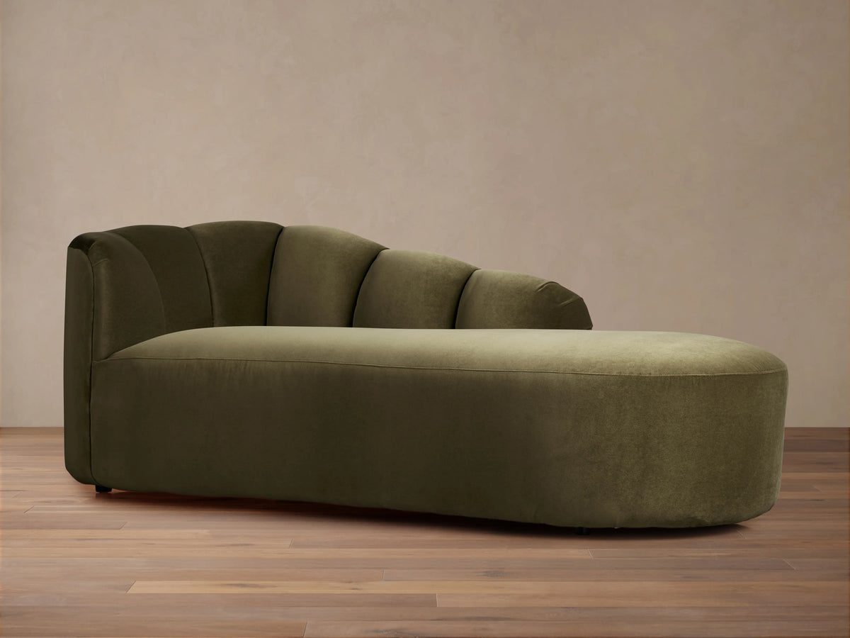 Amira Daybed, an olive green velvet chaise lounge with a channel-tufted backrest & rounded edges, on a wooden floor.