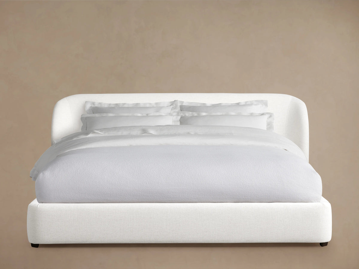 Adalynn Bed with upholstered white headboard, side view. Light gray bedding drapes casually, set against a neutral backdrop.