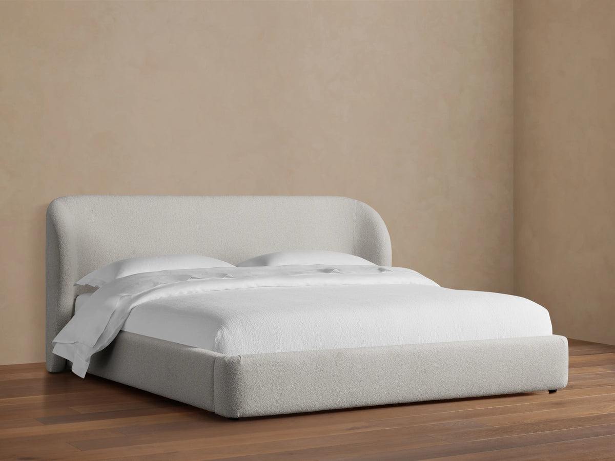 Adalynn Bed with a curved upholstered headboard and low-profile frame, in light beige fabric, set on wooden floor.