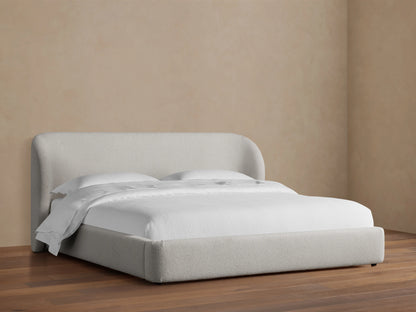 Adalynn Bed with a curved upholstered headboard and low-profile frame, in light beige fabric, set on wooden floor.