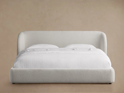 Adalynn Bed with a curved upholstered headboard in light beige fabric, neatly made with white bedding, on a wooden floor.