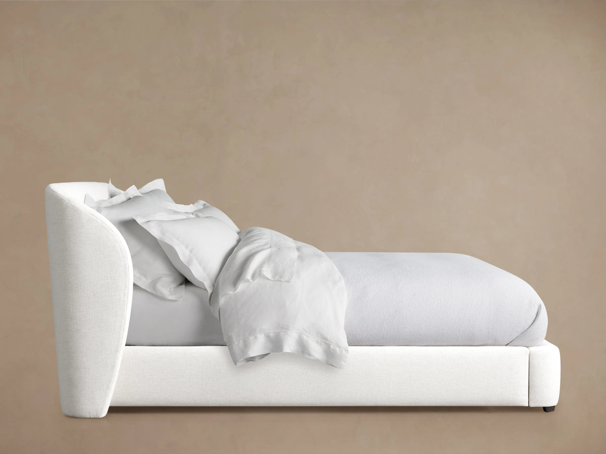 Adalynn Bed in side view with an upholstered white headboard and base. It has wrinkled white bedding and sits against a beige background.