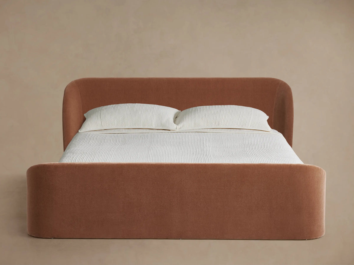 Adalynn Bed with high, rounded footboard and curved headboard, upholstered in soft rust fabric, white bedding.
