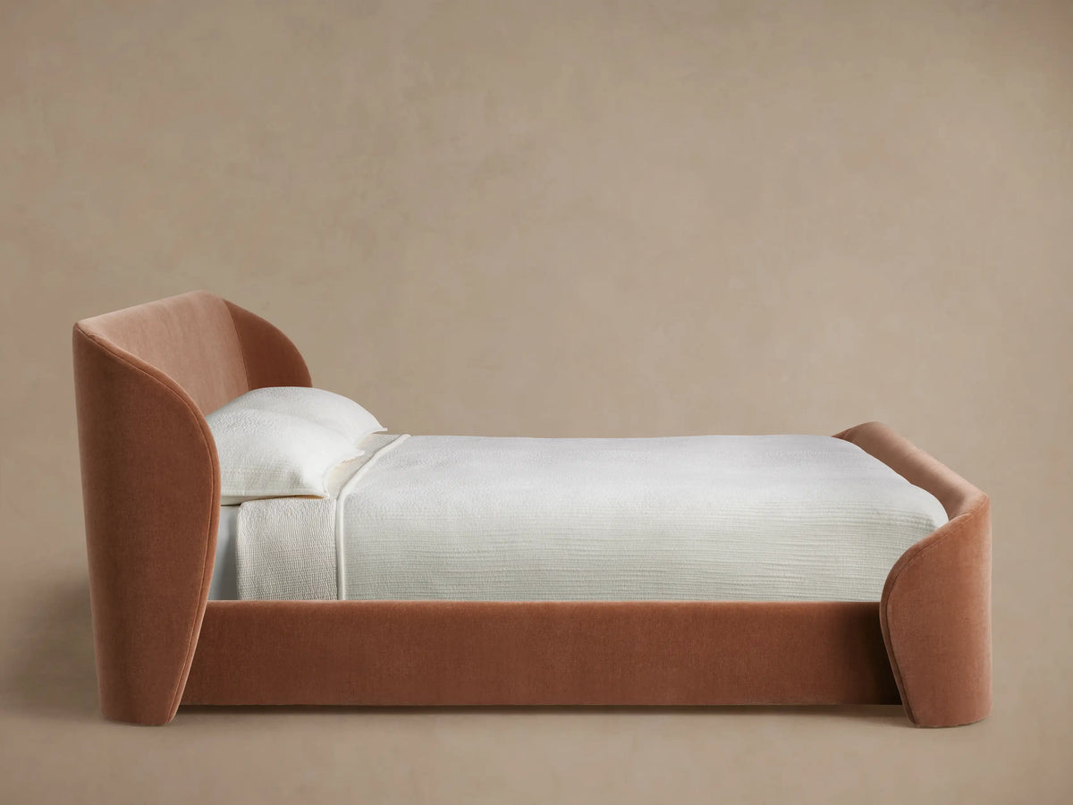 Close-up of Adalynn Bed's upholstered headboard: a soft, curved reddish-brown fabric edge against a light wall.