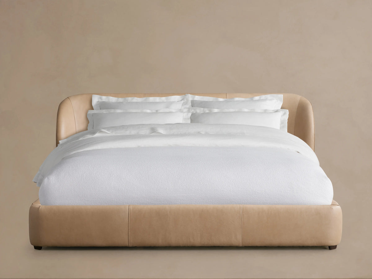 Adalynn Leather Bed with a curved, padded leather headboard in a light tan shade, dressed with white bedding, against a neutral background.