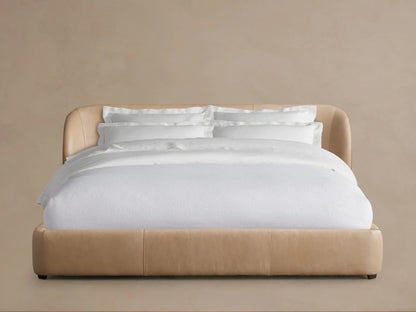Adalynn Leather Bed with a curved, padded leather headboard in a light tan shade, dressed with white bedding, against a neutral background.