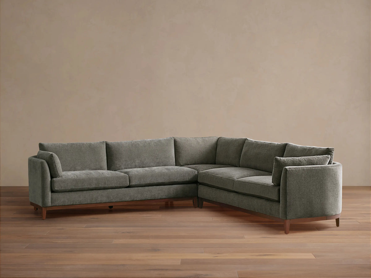 Bainbridge Three-Piece Corner Sectional, L-shaped, in deep moss green fabric, with exposed wood base & legs, on a wood floor.