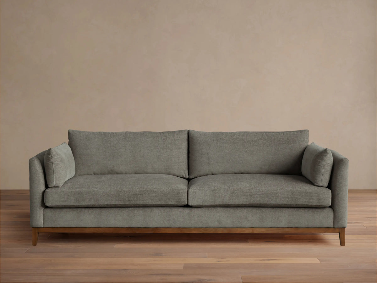 Grey fabric Bainbridge Sofa with two seat & back cushions, bolster arm pillows, and a light wood base on a wood floor.