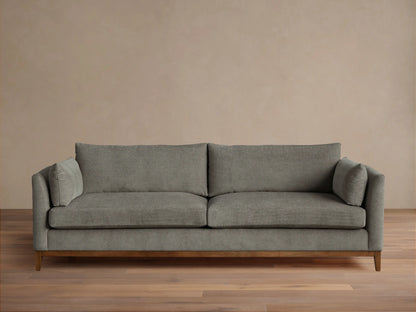 Grey fabric Bainbridge Sofa with two seat & back cushions, bolster arm pillows, and a light wood base on a wood floor.