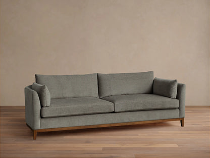 Grey fabric Bainbridge Sofa with two seat & back cushions, bolster arm pillows, and a light wood base on a wood floor.