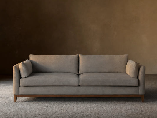 Grey fabric Bainbridge Sofa with two seat & back cushions, bolster arm pillows, and a light wood base on a wood floor.