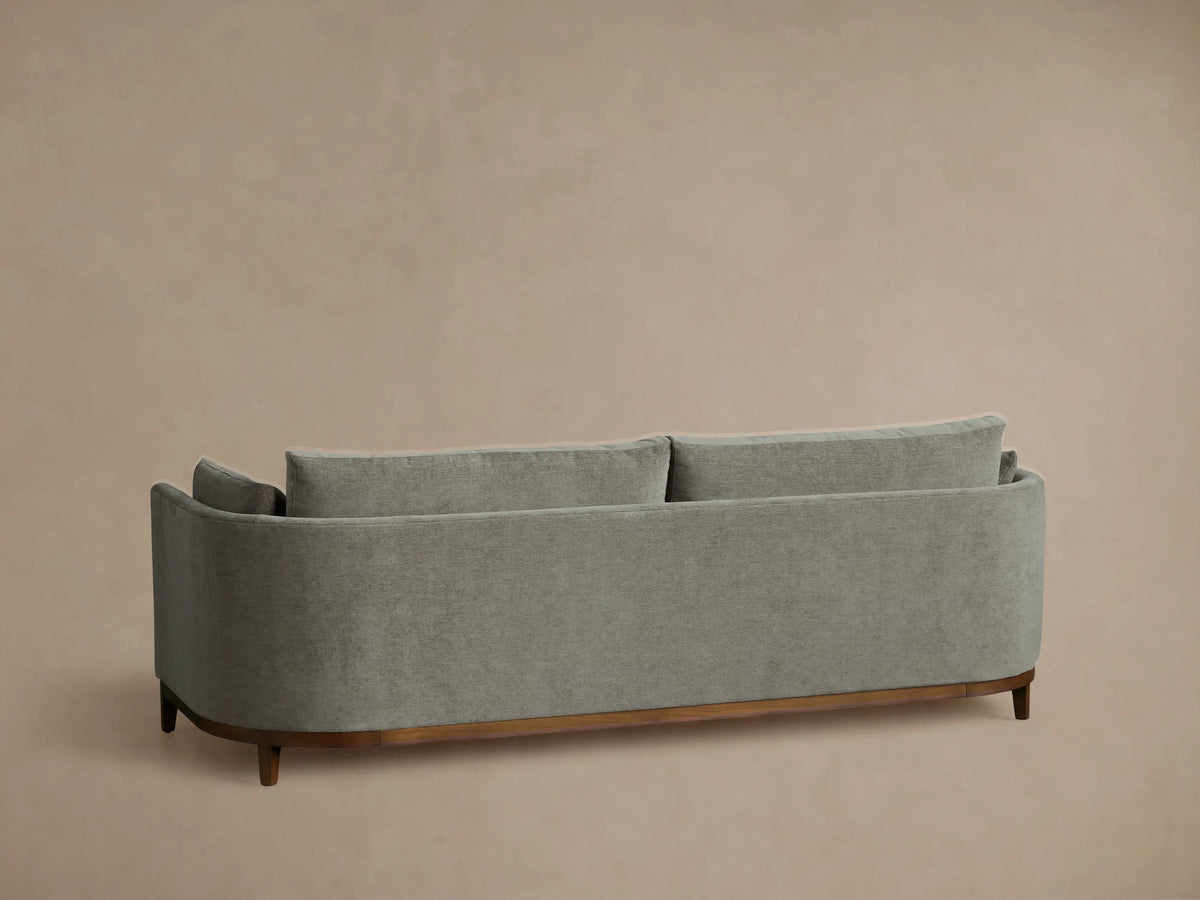 Angled close-up of the Bainbridge Sofa, featuring textured gray fabric, curved armrest with cushion, and dark wooden base.