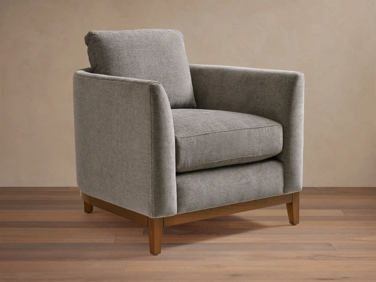 Gray upholstered armchair with square design, featuring plush seat and back cushions. Wooden legs on a hardwood floor, neutral background.