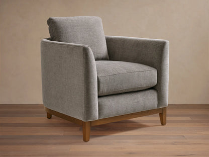Gray upholstered armchair with square design, featuring plush seat and back cushions. Wooden legs on a hardwood floor, neutral background.