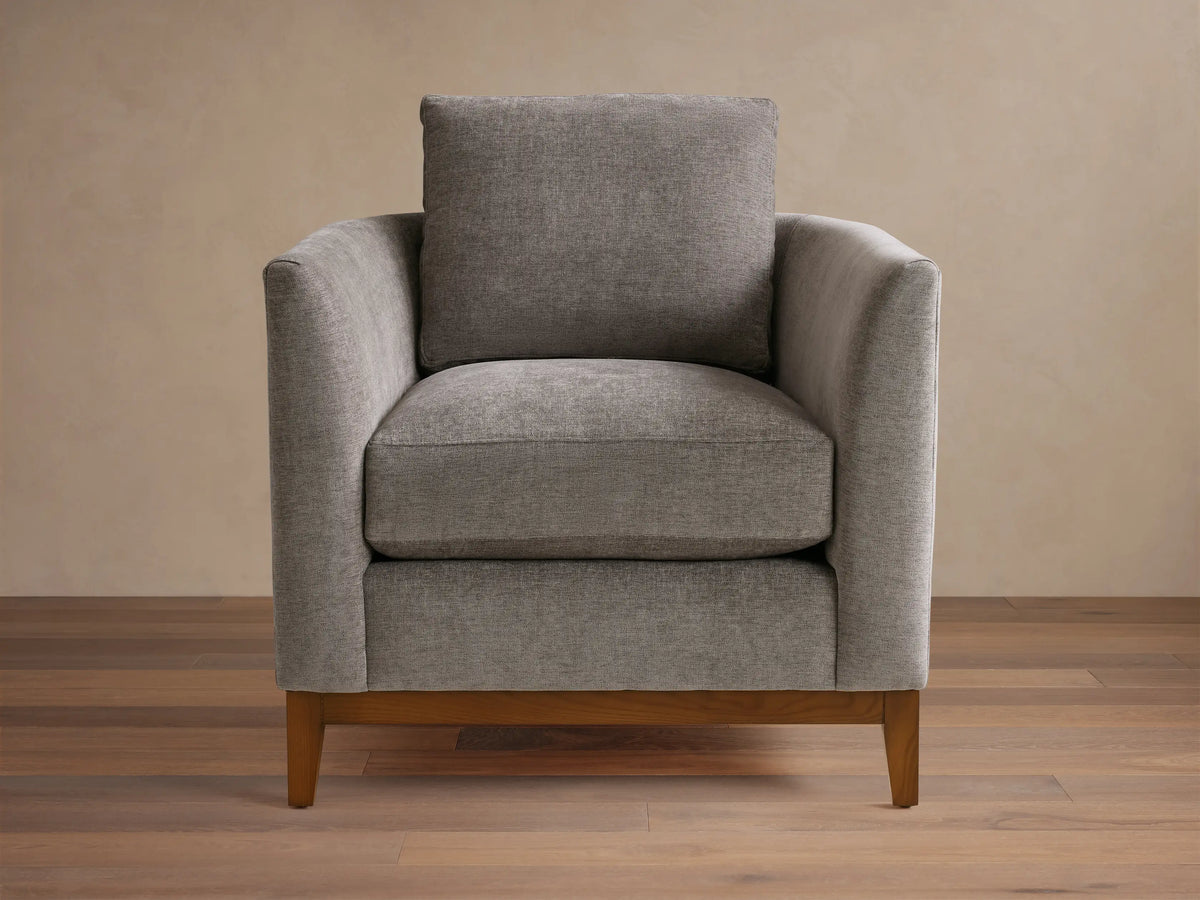 Gray upholstered armchair with square design, featuring plush seat and back cushions. Wooden legs on a hardwood floor, neutral background.