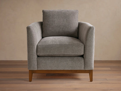 Gray upholstered armchair with square design, featuring plush seat and back cushions. Wooden legs on a hardwood floor, neutral background.