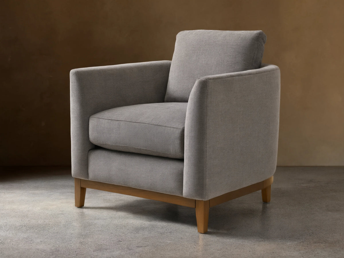 Gray upholstered armchair with square design, featuring plush seat and back cushions. Wooden legs on a hardwood floor, neutral background.