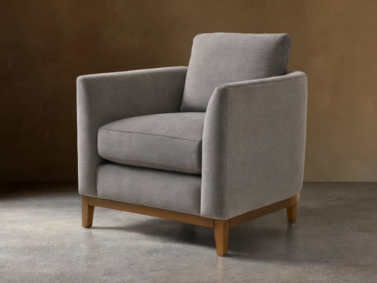 Gray upholstered armchair with square design, featuring plush seat and back cushions. Wooden legs on a hardwood floor, neutral background.