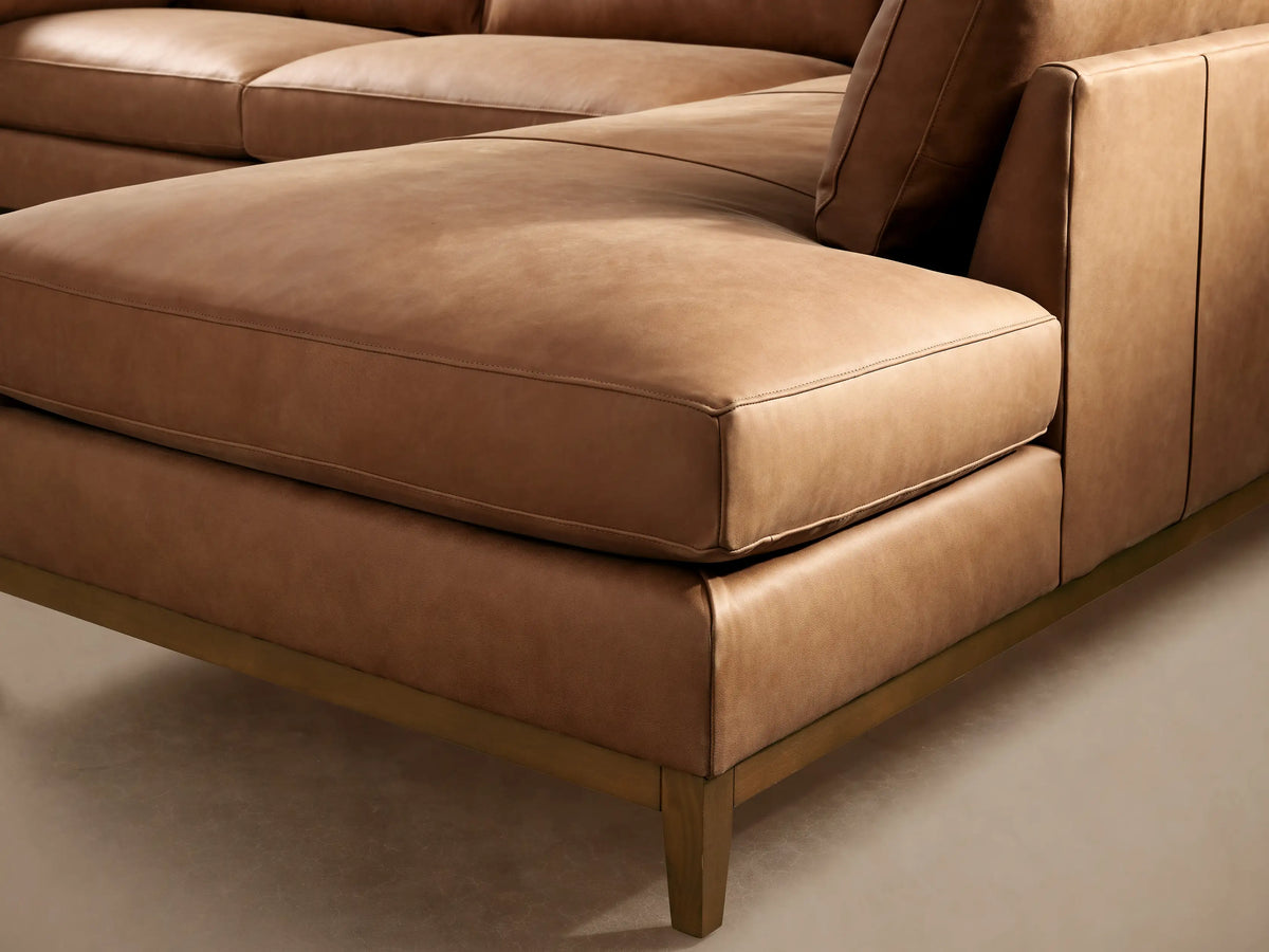 Tan leather Bainbridge Two-Piece Bumper Sectional, L-shaped, with a light wood frame, on a wooden floor.