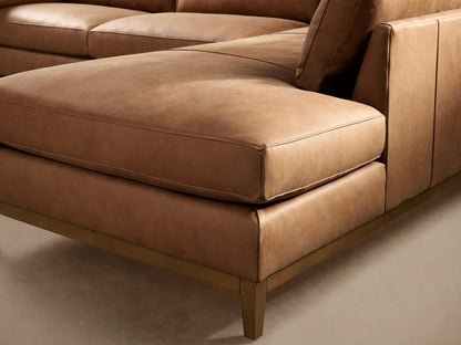 Tan leather Bainbridge Two-Piece Bumper Sectional, L-shaped, with a light wood frame, on a wooden floor.