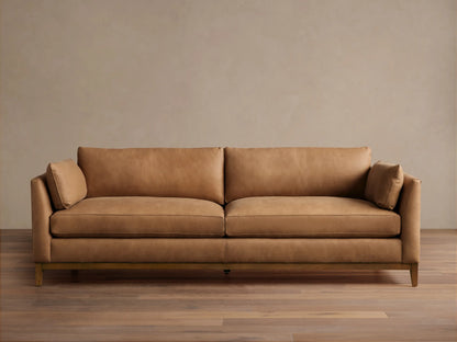 Smooth tan leather Bainbridge sofa with two seat cushions, two back cushions, and small arm pillows on a visible wooden base.