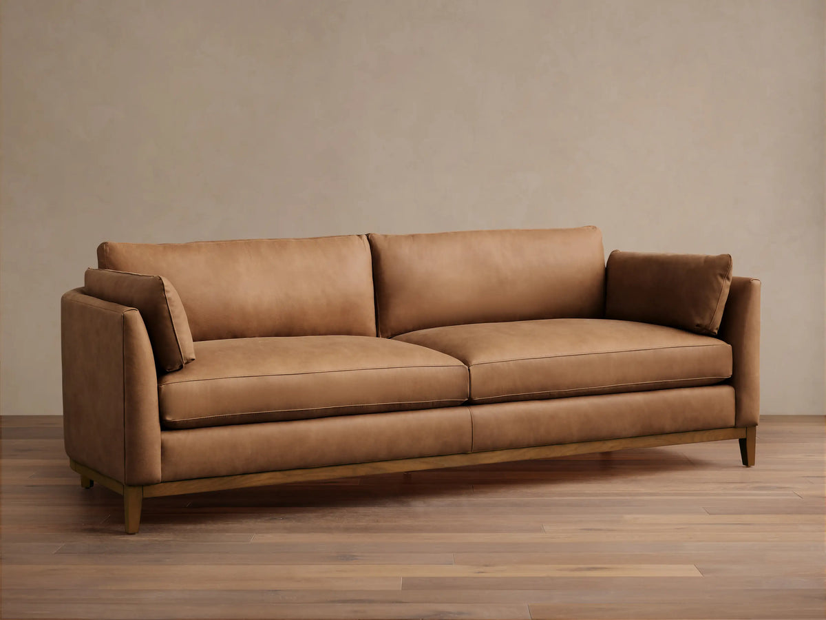 Smooth tan leather Bainbridge sofa with two seat cushions, two back cushions, and small arm pillows on a visible wooden base.