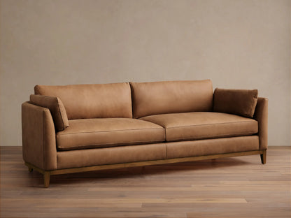 Smooth tan leather Bainbridge sofa with two seat cushions, two back cushions, and small arm pillows on a visible wooden base.