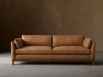 Smooth tan leather Bainbridge sofa with two seat cushions, two back cushions, and small arm pillows on a visible wooden base.