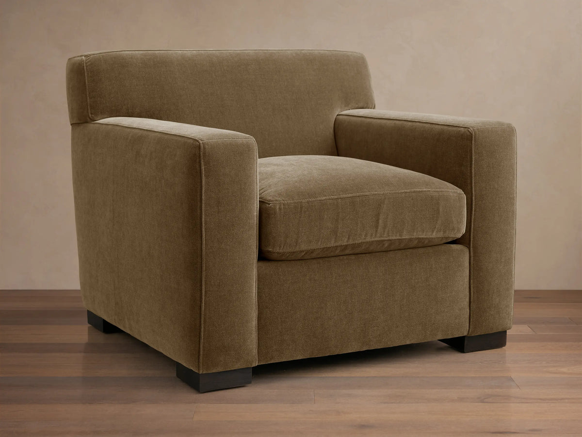 Bronte Chair upholstered in muted olive-brown fabric, angled view, on a wooden floor. Modern, boxy design.