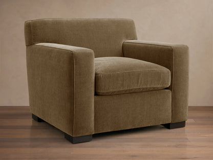 Bronte Chair upholstered in muted olive-brown fabric, angled view, on a wooden floor. Modern, boxy design.
