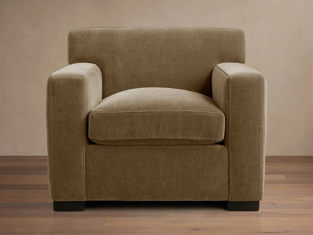 Bronte Chair upholstered in muted olive-brown fabric, angled view, on a wooden floor. Modern, boxy design.