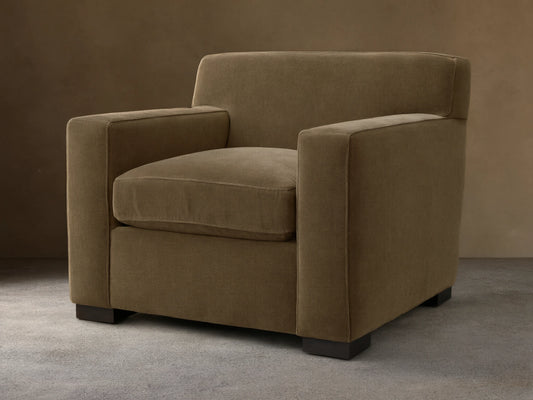 Bronte Chair upholstered in muted olive-brown fabric, angled view, on a wooden floor. Modern, boxy design.