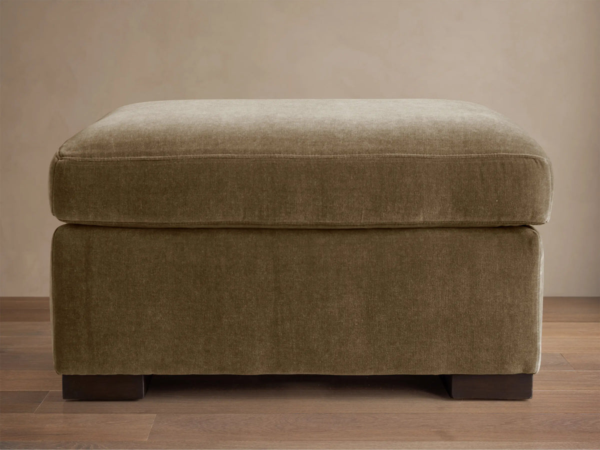 Bronte Ottoman: plush, rectangular, muted olive green fabric. Soft top cushion, dark block feet on a wood floor.