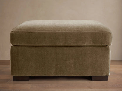 Bronte Ottoman: plush, rectangular, muted olive green fabric. Soft top cushion, dark block feet on a wood floor.