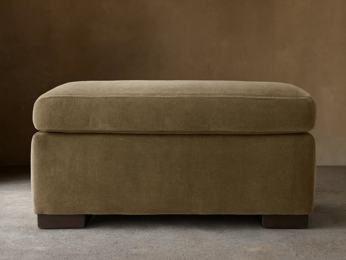Bronte Ottoman: plush, rectangular, muted olive green fabric. Soft top cushion, dark block feet on a wood floor.