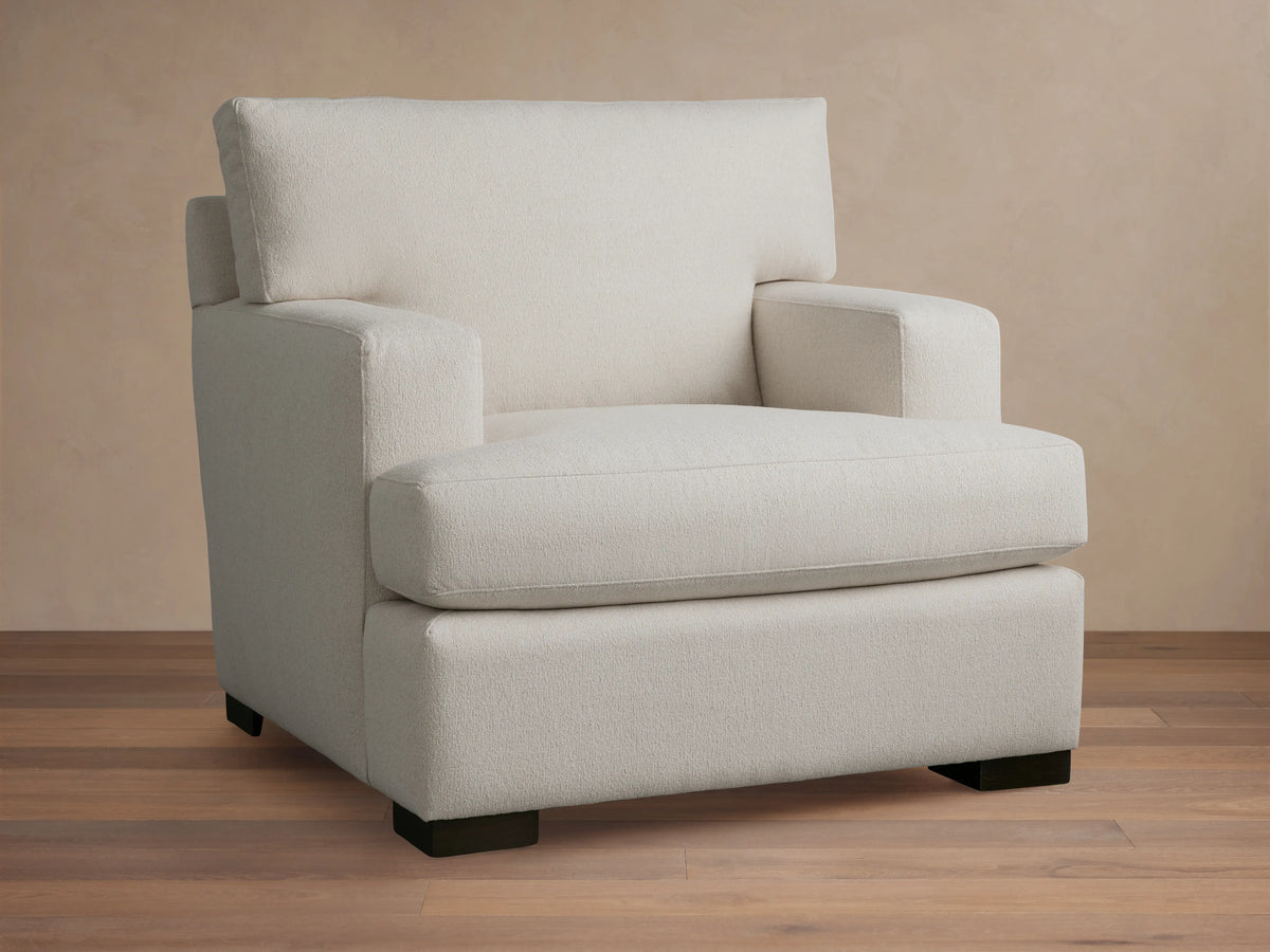 White Dune Chair with square arms and cushioned seat and backrest. Features a minimalistic design with dark wooden legs on a wooden floor.