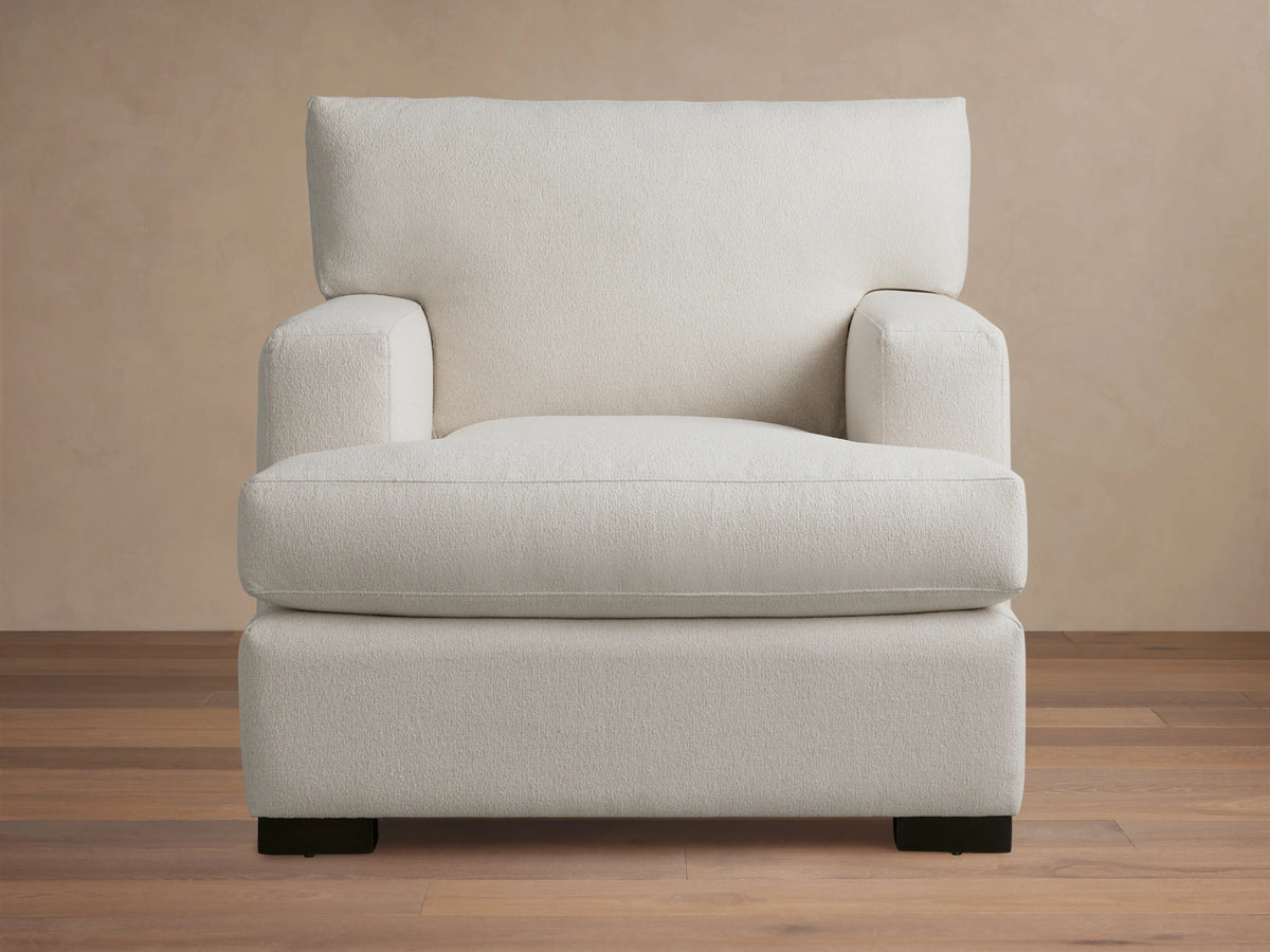 White Dune Chair with square arms and cushioned seat and backrest. Features a minimalistic design with dark wooden legs on a wooden floor.
