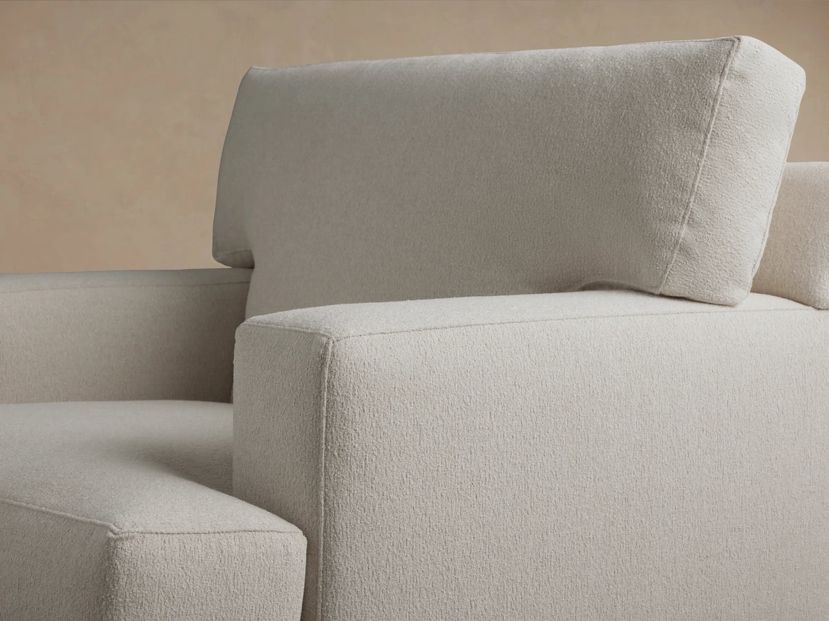 Front view of the Dune Chair featuring a soft beige fabric, plush backrest, and wide, cushioned seat. It has straight wooden legs and padded armrests on a wooden floor.