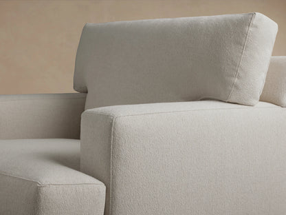 Front view of the Dune Chair featuring a soft beige fabric, plush backrest, and wide, cushioned seat. It has straight wooden legs and padded armrests on a wooden floor.