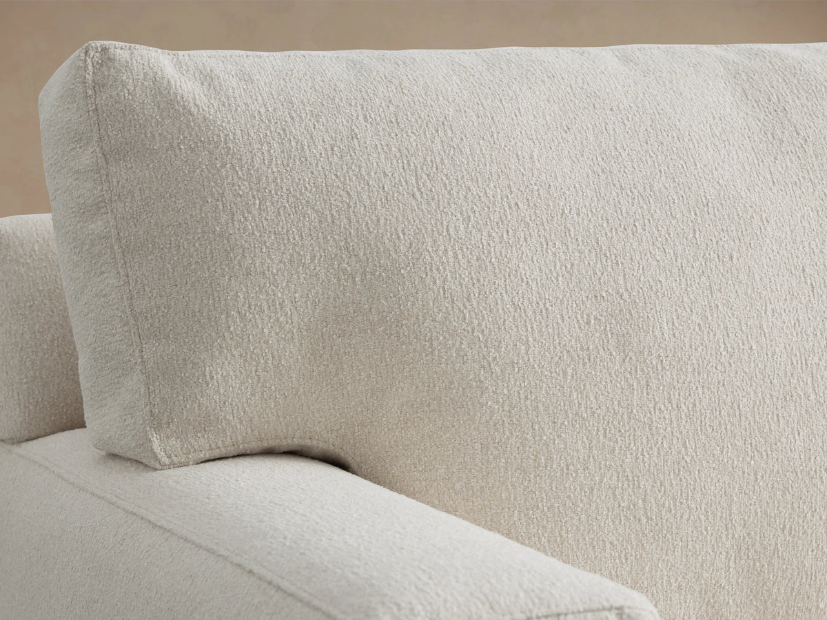 Dune Sofa in light beige fabric, featuring plush back cushions and cushioned seats. Boxy arms and dark wooden legs on a wooden floor.