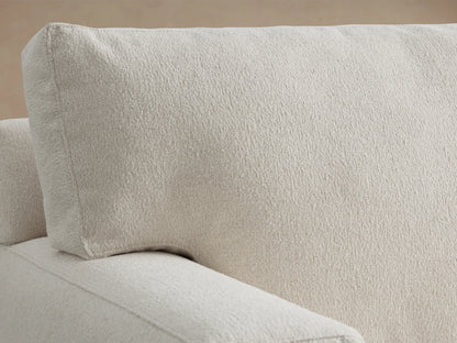 Dune Sofa in light beige fabric, featuring plush back cushions and cushioned seats. Boxy arms and dark wooden legs on a wooden floor.