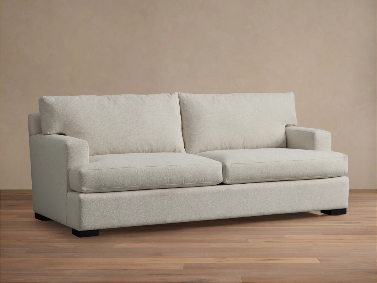A cream-colored Dune Sofa with plush cushions and armrests, set against a beige wall on a wooden floor, captured from the front.