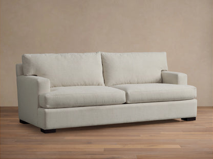 A cream-colored Dune Sofa with plush cushions and armrests, set against a beige wall on a wooden floor, captured from the front.