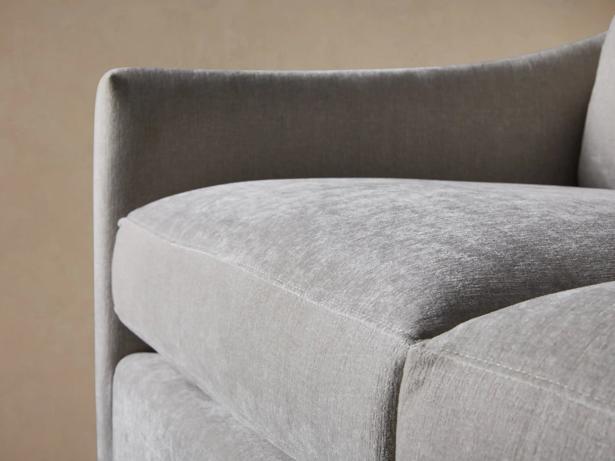 Branson Sofa close-up: light gray plush fabric, rounded armrest, and soft back cushions against a tan wall.