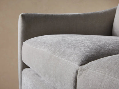 Branson Sofa close-up: light gray plush fabric, rounded armrest, and soft back cushions against a tan wall.