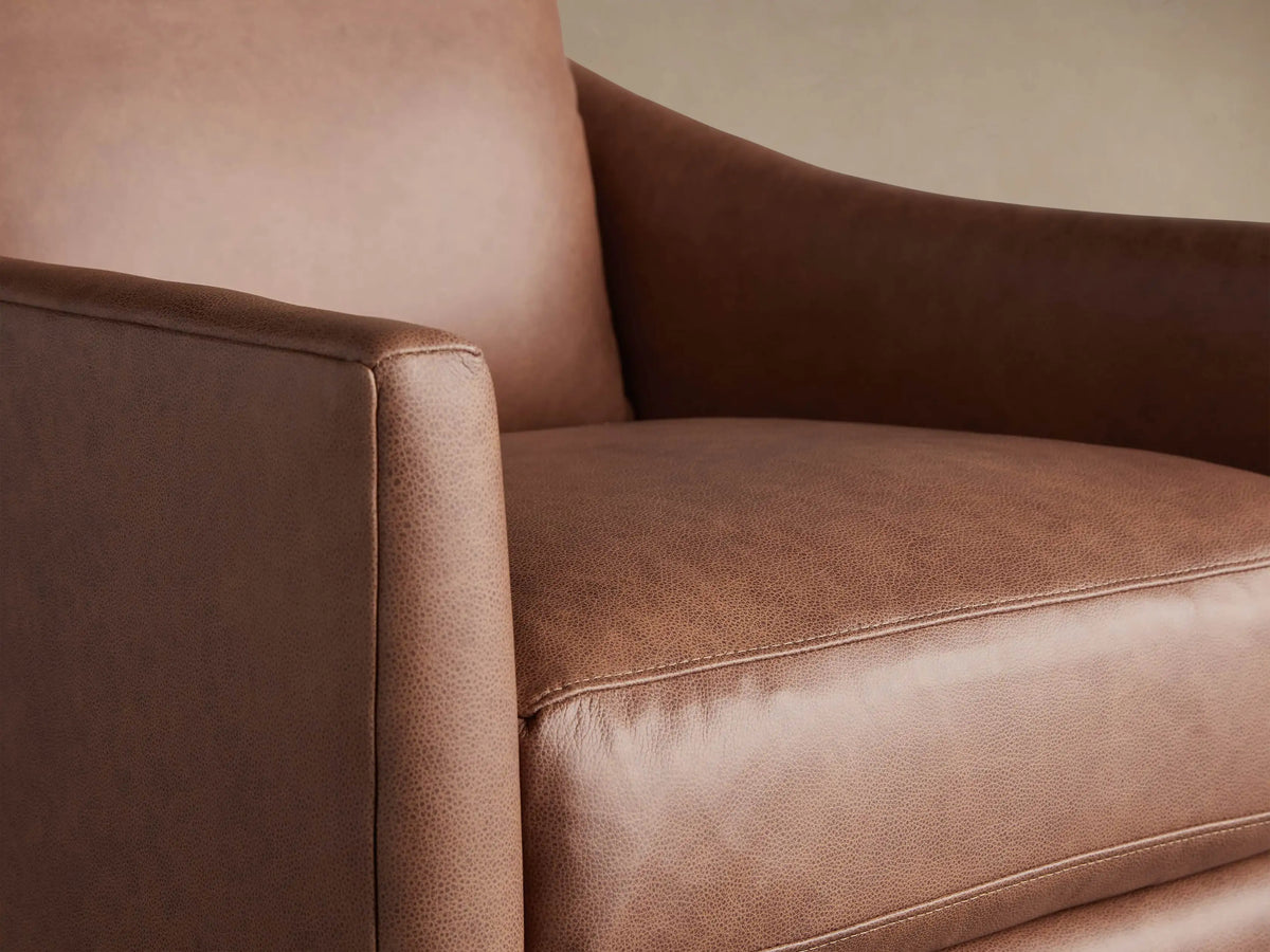 A plush cognac brown leather swivel chair with thick cushions and wide arms, against a warm wall and wood floor.