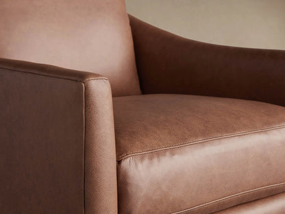 A plush cognac brown leather swivel chair with thick cushions and wide arms, against a warm wall and wood floor.