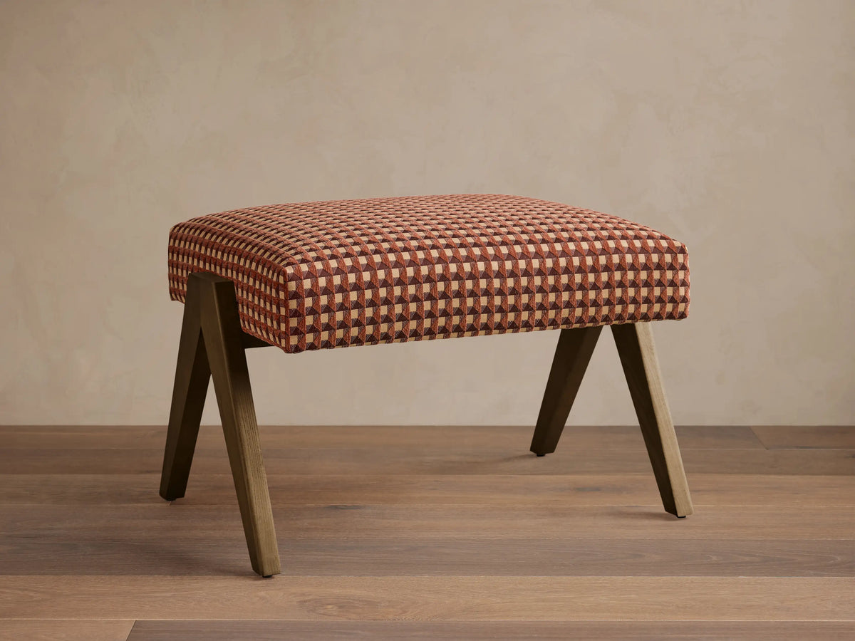 Front view of the Elias Ottoman with red, brown, and beige geometric patterned upholstery and angled wooden legs.