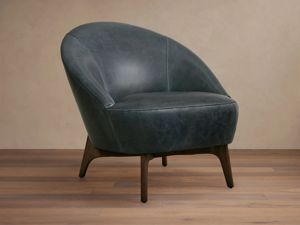 Elodie Leather Chair in dark grey leather with a rounded backrest and wooden legs, on a wooden floor with a beige wall backdrop.